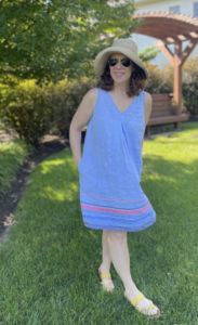 white woman in a denim colored shift dress with pink detailing towards the bottom of the dress with her hands in her pockets wearing sunglasses and a sunhat