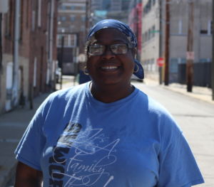 older black woman wearing a sky blue t-shirt, a blue bandana and glasses smiling with teeth