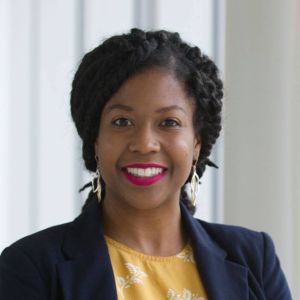 black woman wearing dark navy blue blazer and yellow blouse with white flowers on it smiling with teeth wearing bright red lipstick