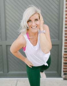 photo of emily nichols wearing a white tank top with green pants and she is smiling with teeth in front of a garage