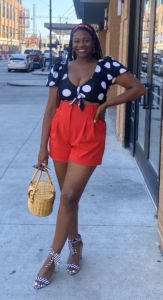 a photo of erica scott wearing red shorts and a blue and white polka dot blouse standing and smiling with her hand on her hip 