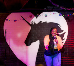 photo of tocarra mallard wearing jeans, black blouse and orange jacket doing standup comedy at a comedy club