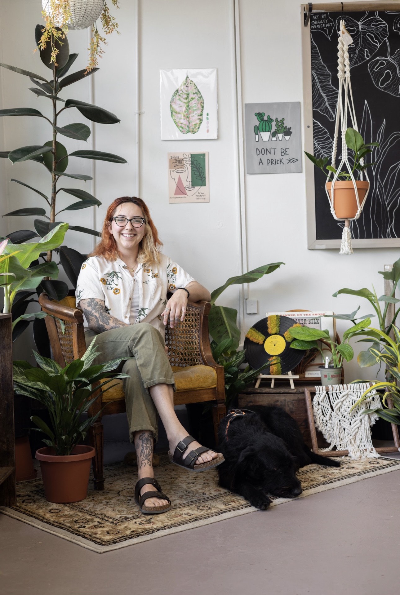Hannah Flege sitting in a chair in Urban Houseplant Collective with a black dog laying by her feet.