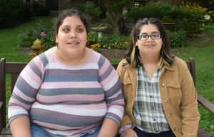 Two visually impaired young women, Samantha (left) and Nikki (right), are featured from the waist up, smiling, wearing casual clothing, and seated next to one another on a bench in a garden.