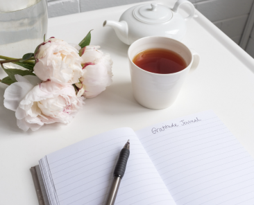 Open journal with writing that reads "gratitude journal" a cup of coffee and some flowers sit nearby