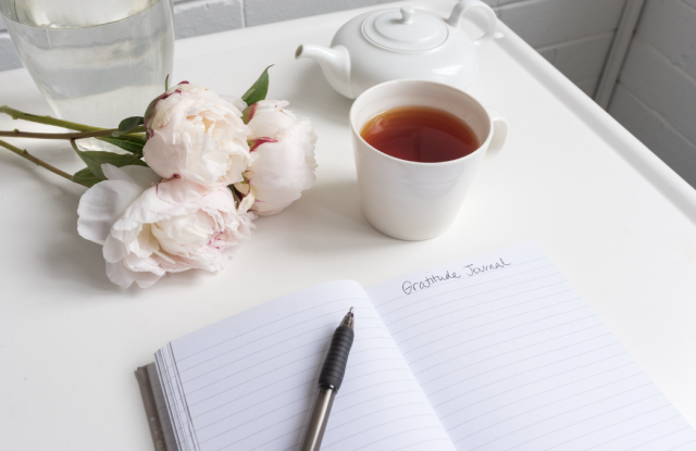 Open journal with writing that reads "gratitude journal" a cup of coffee and some flowers sit nearby