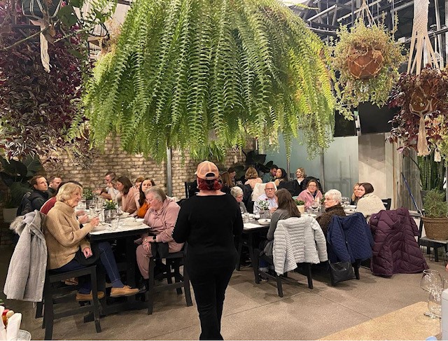 Sully's Grill dinning room with people sitting down. One woman standing. Plants hanging from the ceiling.