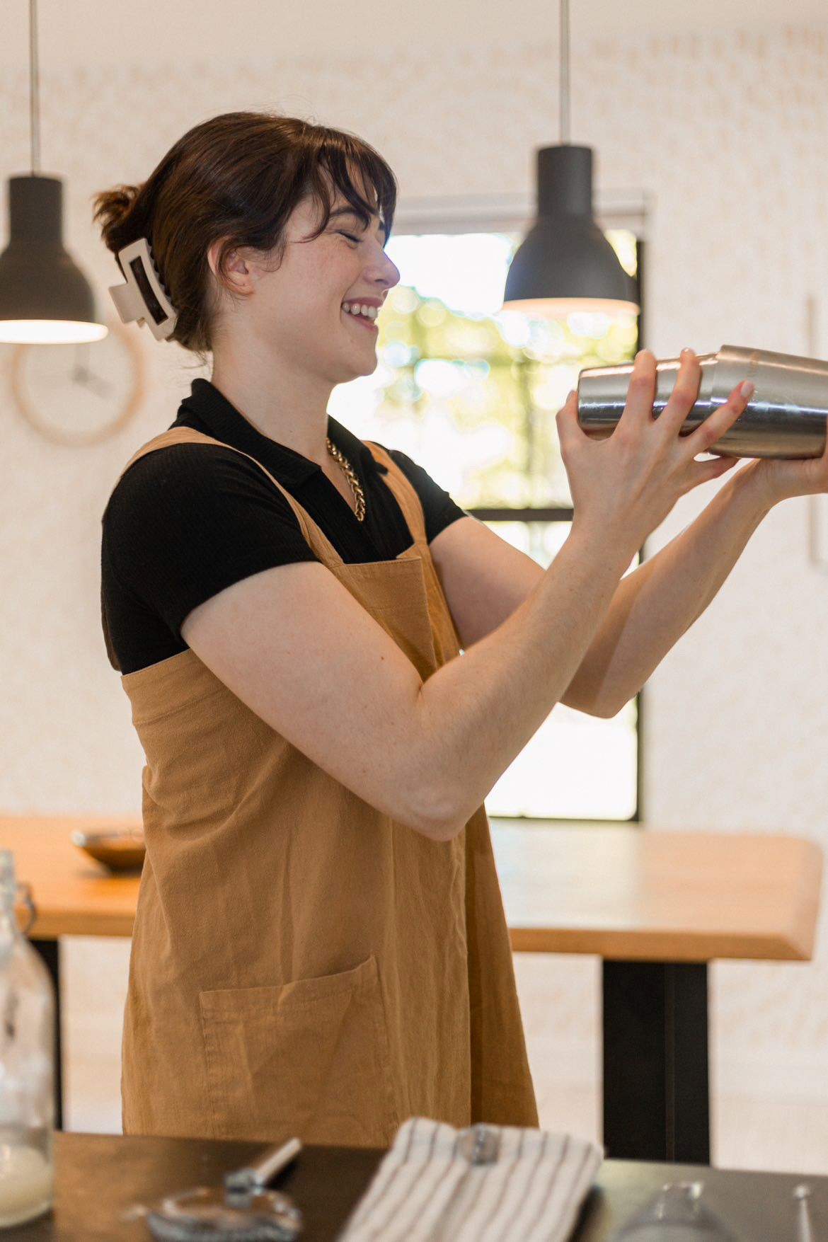 A woman in an apron smiling while making a drink in a shaker.