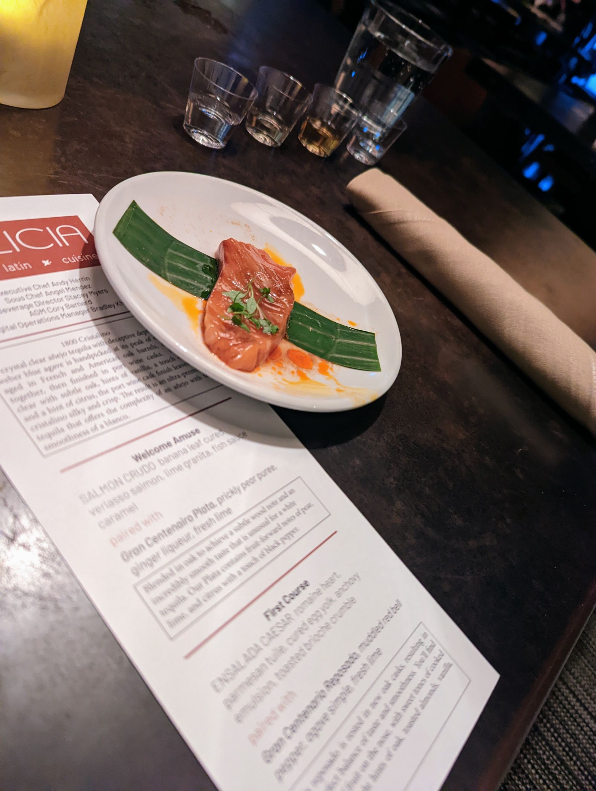 4c Menu set underneath an plate of food from Dalicia's. Silverwear wrapped in beige cloth and shot glasses in the background.