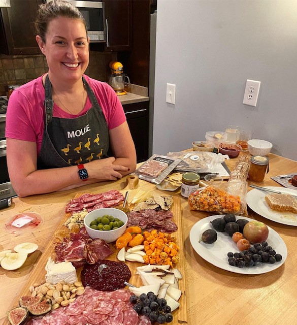 Woman smiles at the camera with a charcuterie platter in front of her.