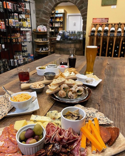 Charcuterie platter and fixings displayed on a wooden table with wine a beer in the background.