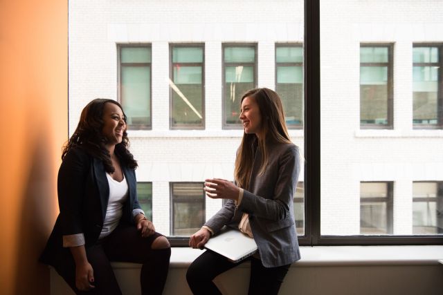 Two women sit on a windowsill laughing
