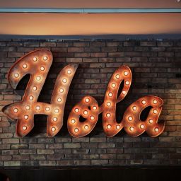 Large orang/rust color neon sign reads "Hola" against a brick wall.