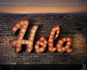 Large orang/rust color neon sign reads "Hola" against a brick wall.