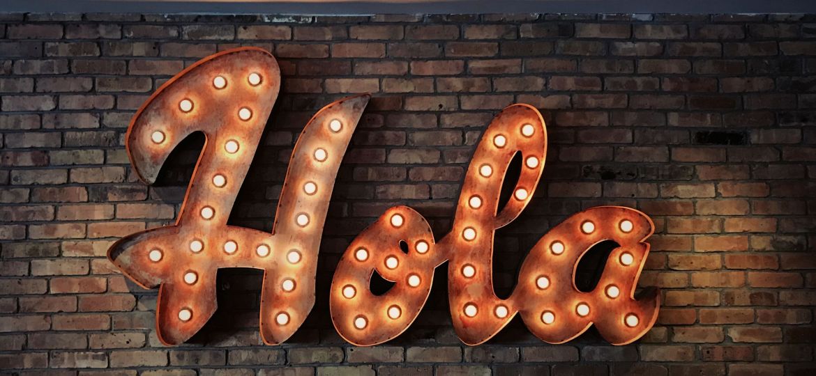 Large orang/rust color neon sign reads "Hola" against a brick wall.