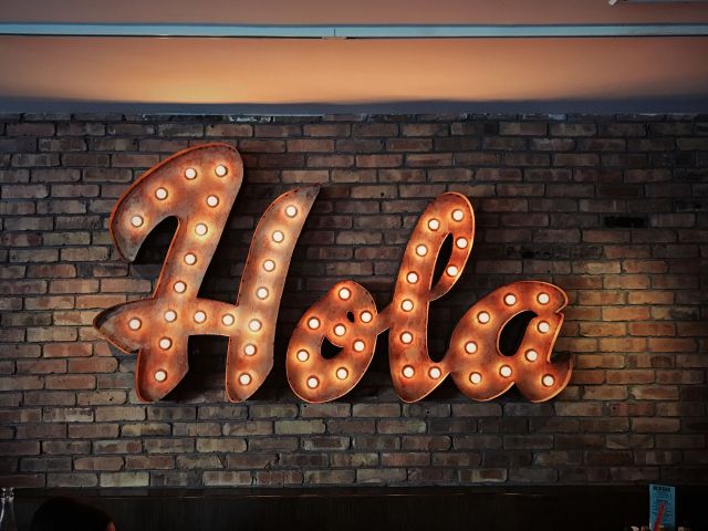 Large orang/rust color neon sign reads "Hola" against a brick wall.