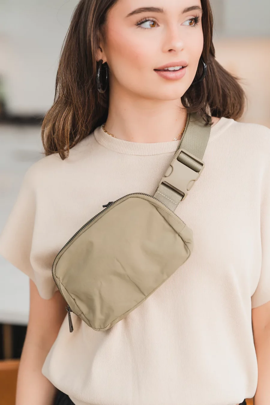 Woman with dark brown hair in a light pink shirt modeling beige fanny pack across her body