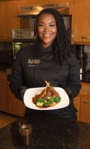 Jazymn Perry holding a plate of food in the kitchen. She has dark hair and is dressed in a chef's shirt