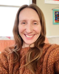 Lauren Hall Bushman wearing a orange sweater with long brown wavy hair smiling 