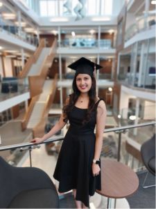 Aishariya B. Alvarez wearing a black dress and graduation cap smiling at the camera in Butler's business building 