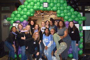 Fourteen women pose in front of a green balloon arch at the 2023 Women Build Kickoff Committee and Homeowners