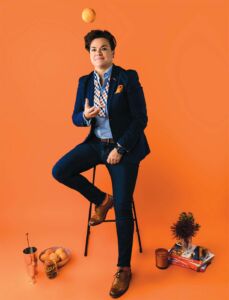 Leanna Chroman sitting in a stool against an orange background throwing an orange in the air