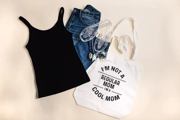I’m a Cool Mom Graphic Tote Bag laying next to jeans and a black tank top