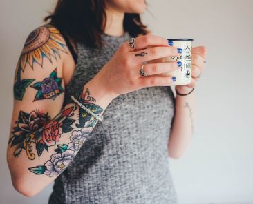 A woman holds a cup of coffee and sports a sleeve tat on her arm.
