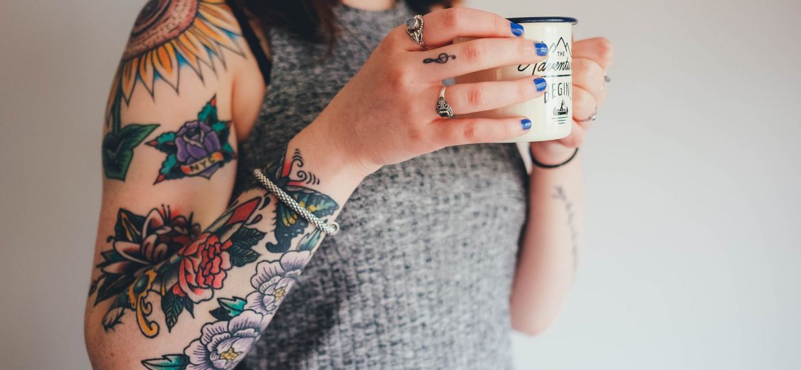 A woman holds a cup of coffee and sports a sleeve tat on her arm.