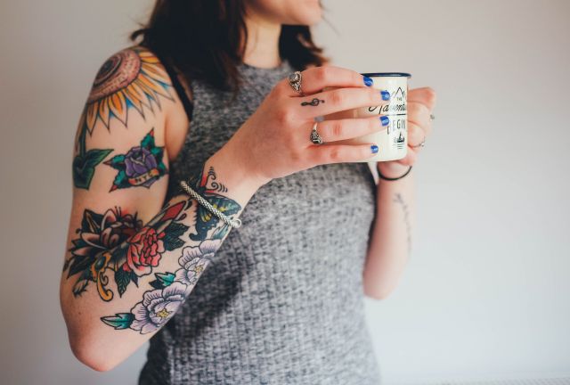 A woman holds a cup of coffee and sports a sleeve tat on her arm.