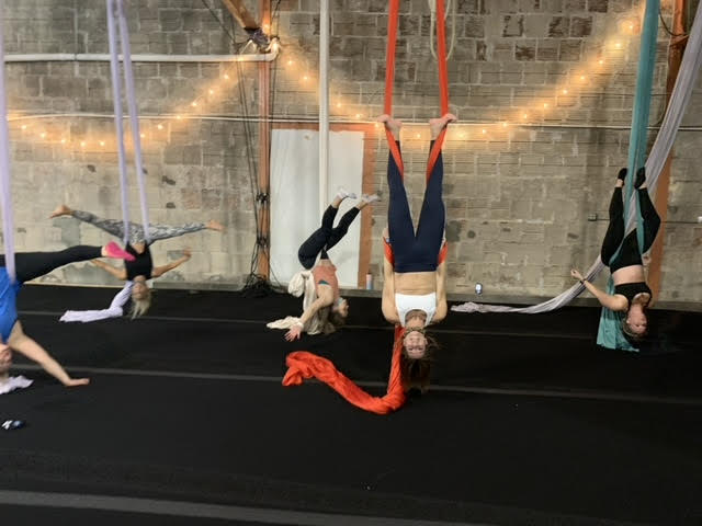 Women doing yoga on silks. A brick wall in the background with string lights hanging from the ceiling.