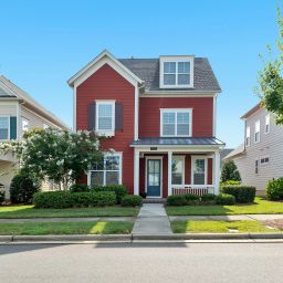 Red house in between two houses on a tree lined street with bright blue sky behind.