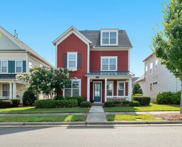 Red house in between two houses on a tree lined street with bright blue sky behind.