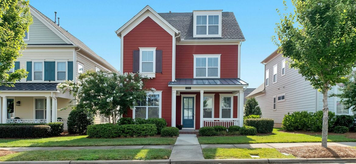 Red house in between two houses on a tree lined street with bright blue sky behind.