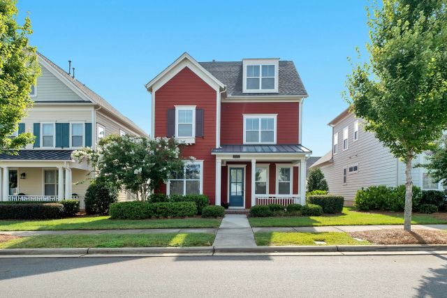 Red house in between two houses on a tree lined street with bright blue sky behind.