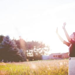 Woman standing in field raising hands in the air, sunlight flare.