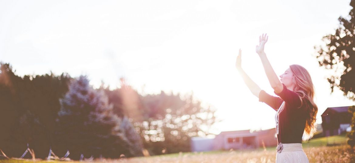 Woman standing in field raising hands in the air, sunlight flare.