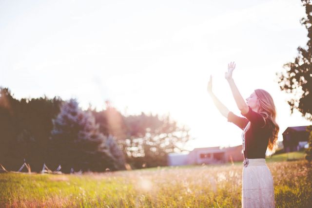 Woman standing in field raising hands in the air, sunlight flare.