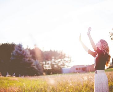 Woman standing in field raising hands in the air, sunlight flare.