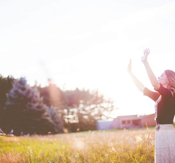 Woman standing in field raising hands in the air, sunlight flare.