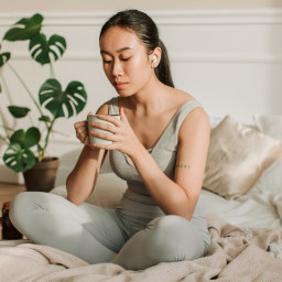 Woman meditating with mug in hand on white bed with blush colored blanket. She has a phonytail and headphones in.