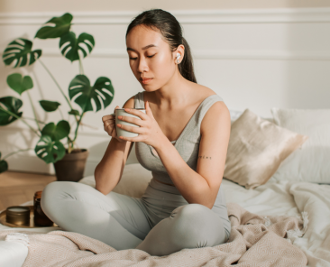 Woman meditating with mug in hand on white bed with blush colored blanket. She has a phonytail and headphones in.