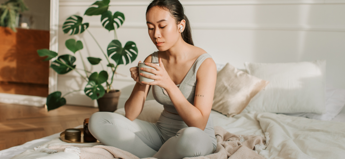 Woman meditating with mug in hand on white bed with blush colored blanket. She has a phonytail and headphones in.