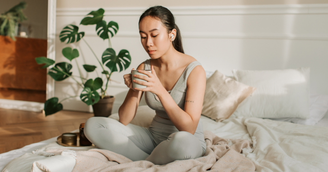 Woman meditating with mug in hand on white bed with blush colored blanket. She has a phonytail and headphones in.