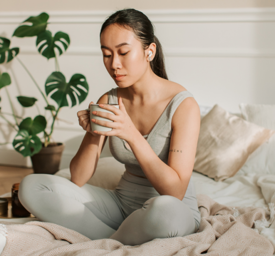 Woman meditating with mug in hand on white bed with blush colored blanket. She has a phonytail and headphones in.