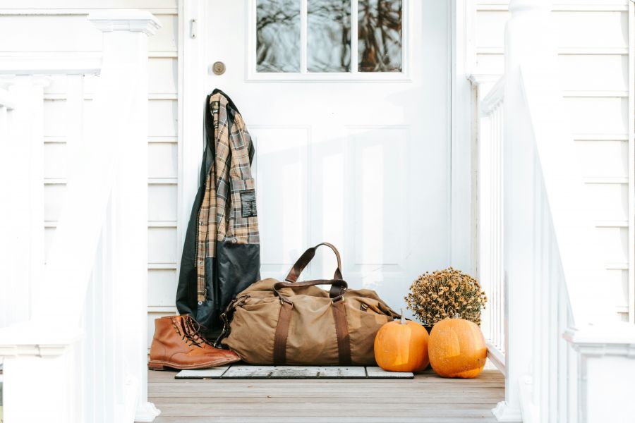 White house, front door with flannel hanging off of the door knob. A duffle bag sits with boots and pumpkins and mums.