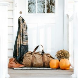 White house, front door with flannel hanging off of the door knob. A duffle bag sits with boots and pumpkins and mums.