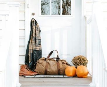 White house, front door with flannel hanging off of the door knob. A duffle bag sits with boots and pumpkins and mums.