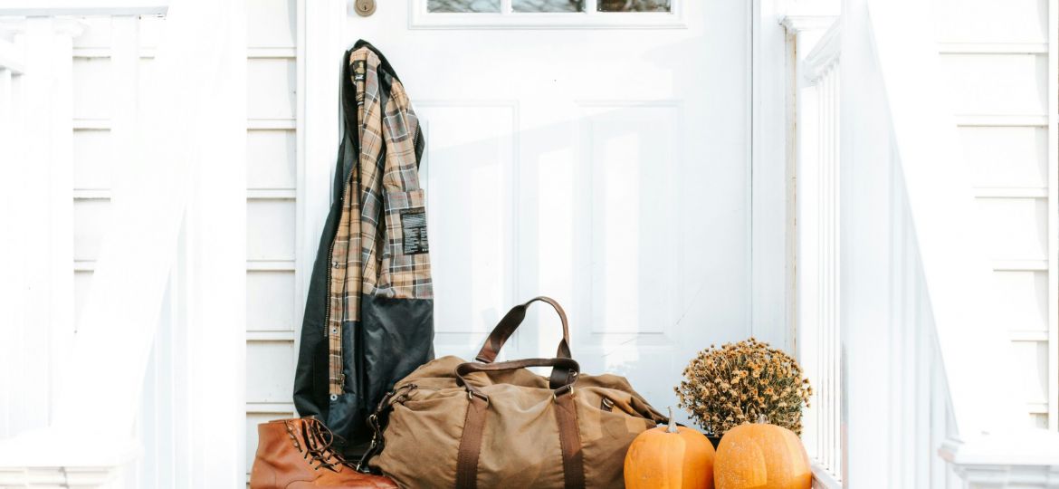 White house, front door with flannel hanging off of the door knob. A duffle bag sits with boots and pumpkins and mums.