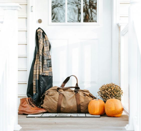 White house, front door with flannel hanging off of the door knob. A duffle bag sits with boots and pumpkins and mums.